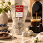 Baboum Momente First-Light-Lungo-Kapseln neben einer Kaffeemaschine, einer Tasse Kaffee, Blaubeeren und Schokolade.