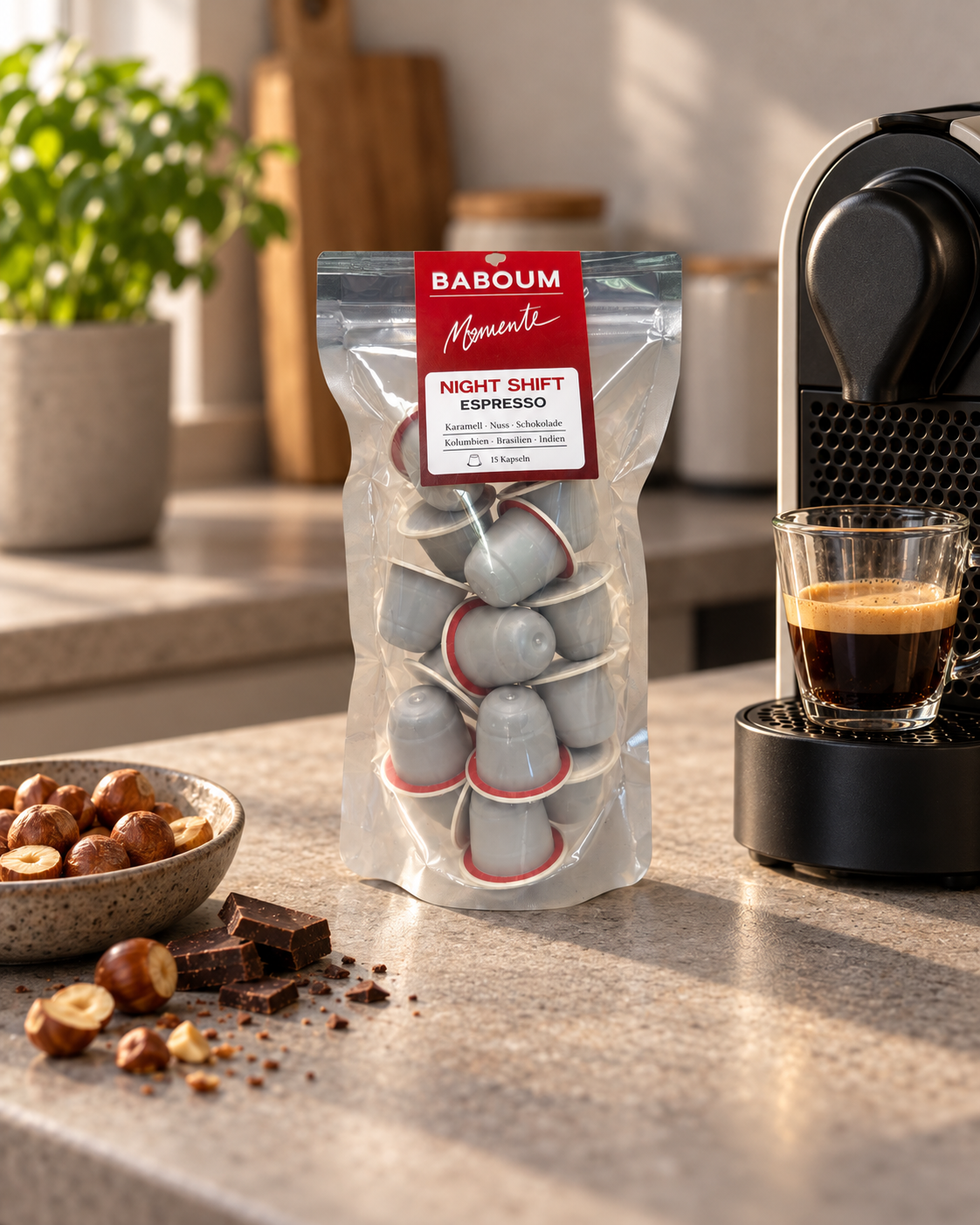 Baboum Momente Night-Shift-Espressokapseln neben einer Kaffeemaschine, einem Espresso, Haselnüssen und Schokolade.